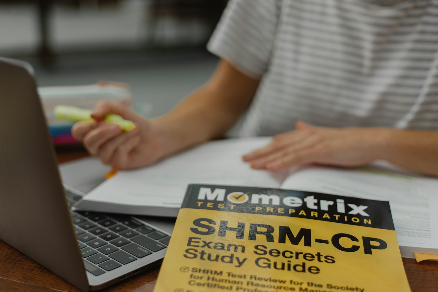 Hands holding a highlighter over a textbook with a laptop.